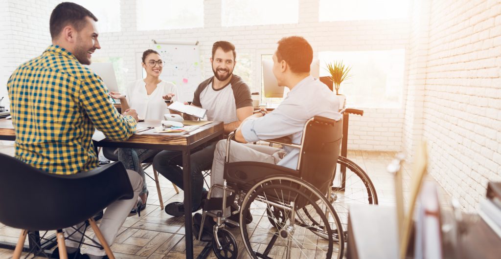 A man in a wheelchair communicates cheerfully with employees of the office during a business meeting.