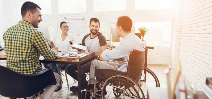 A man in a wheelchair communicates cheerfully with employees of the office during a business meeting.