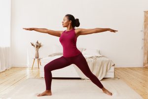 ide view of stylish athletic sporty African girl in leggings and top standing barefooted on floor in warrior 2 pose, doing yoga, strengthening legs, training endurance to get strong healthy body