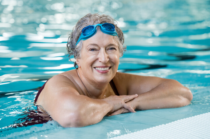 Mature woman wearing swim goggles resting on edge of swimming pool smiling at camera.