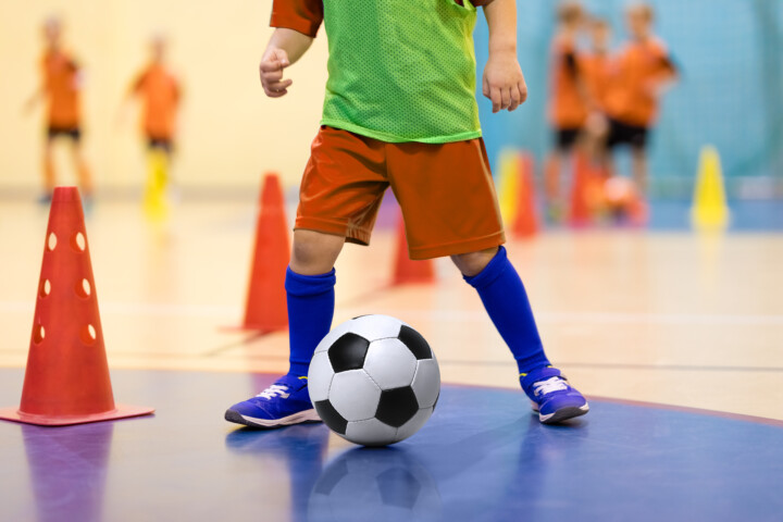 Young soccer player dribbling ball around cones in a gym.