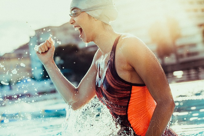 Excited female swimmer with clenched fist celebrating victory in the swimming pool. Woman swimmer cheering success in pool wearing swim goggles and cap.