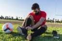 Male soccer player tying cleats and preparing for training outdoors, wearing a mask.