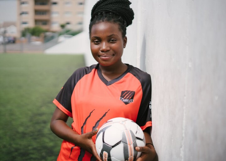 A young BIPOC girl holding a soccer ball leaning on a wall.