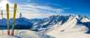 Paire de skis d'hiver posés dans la neige au sommet d'une montagne, avec vue sur une chaîne de montagnes par une journée ensoleillée.