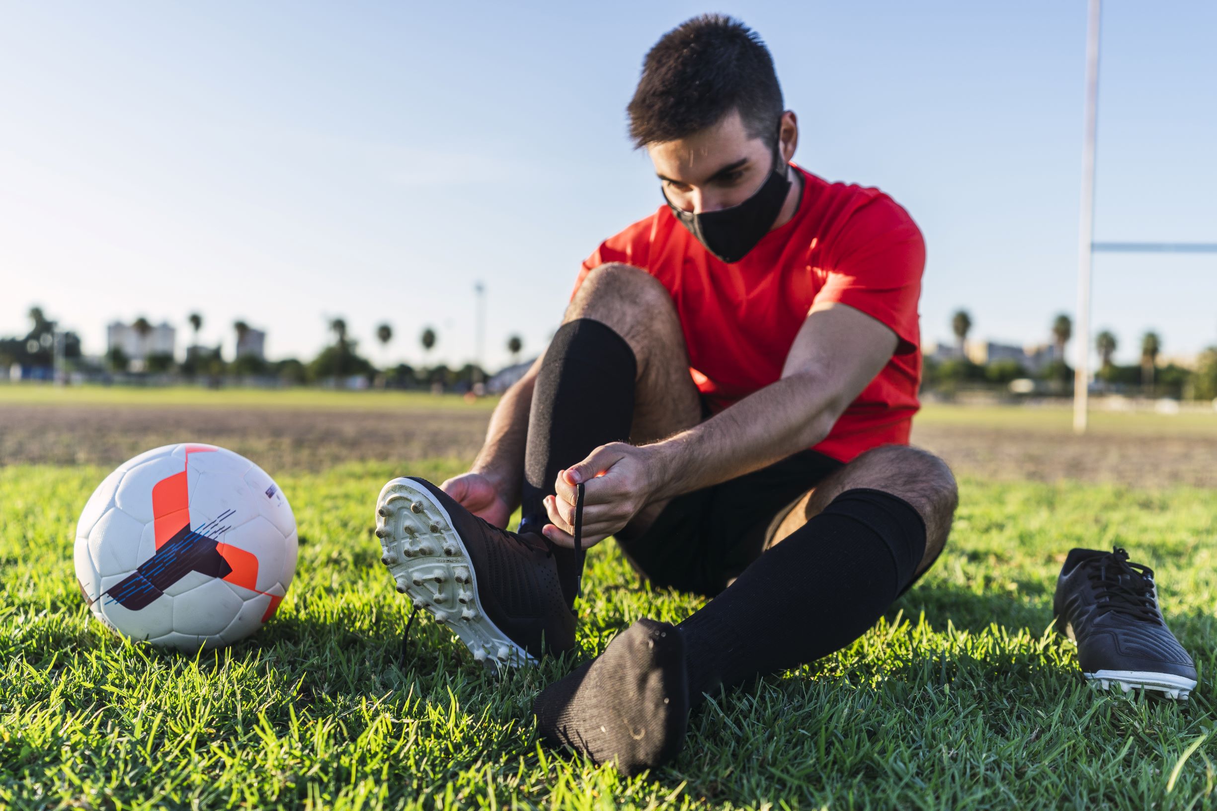 male soccer athlete putting on cleats, preparing to play with mask for covid-19