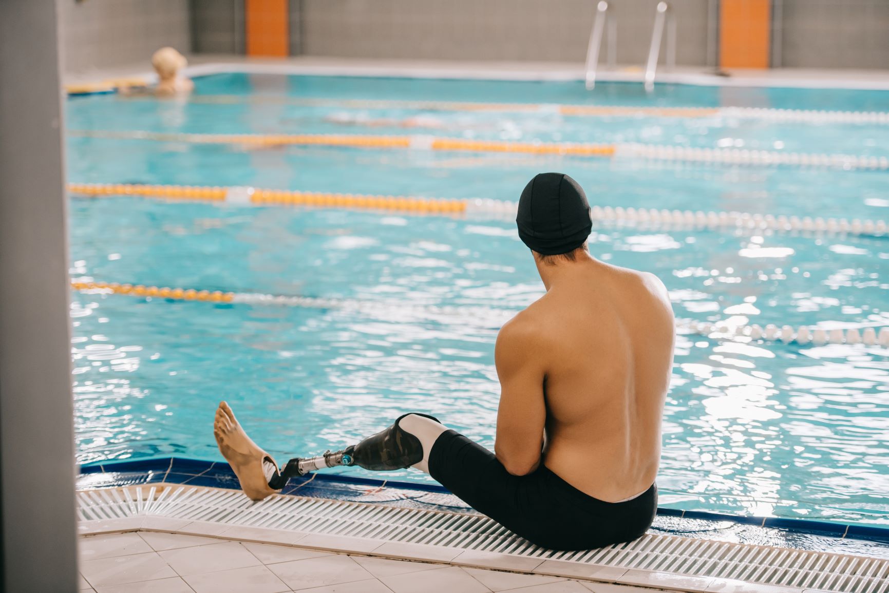 Rear view of young swimmer with artificial leg sitting on poolside of indoor swimming pool