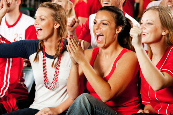 Three women sitting in a stadium, wearing red and cheering on their favourite sports team