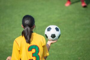 Rear view of a young girl holding a soccer ball on sidelines during a soccer match, wearing a yellow jersey.