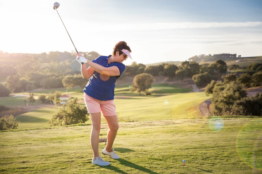 Older adult female playing golf, swinging to hit golf ball on the green.