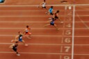 Seven sprinters running across the finish line of a red track.