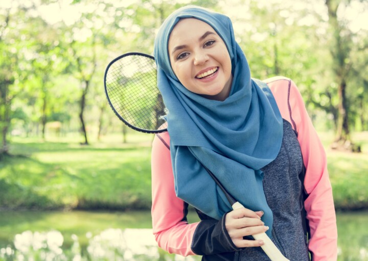 Woman in a park, holding a badminton racquet and smiling at the camera