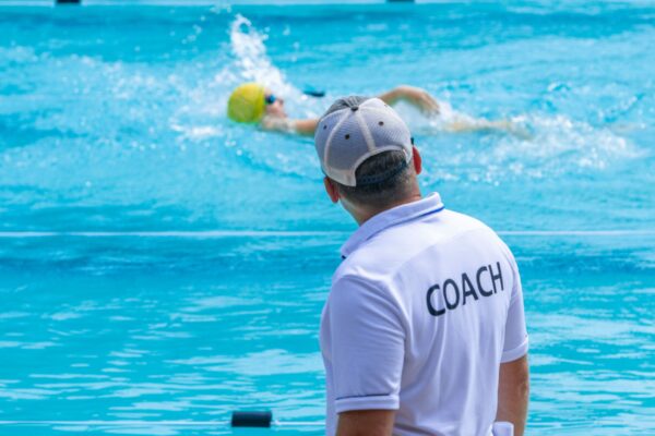 Swimming coach standing beside an outdoor pool and coaching an athlete who is swimming the backstroke.