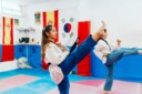 Two girls practicing high kicks in a karate gym with flags in the background.