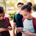 Two school-aged girls on smartphones while walking and wearing backpacks.