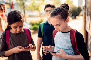 Two school-aged girls on smartphones while walking and wearing backpacks.