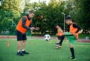 Soccer coach wearing an orange jersey throwing a ball towards a younger player who is kicking the ball back.