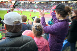 Woman, girl and boy standing applause among fans at a sports stadium, the view from the back