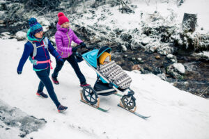 Two women with baby stroller enjoying motherhood in winter forest, mountains landscape. Mother hiking with a partner and a child in white snowy woods. 