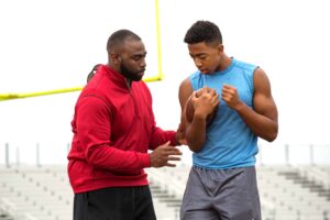 A male football coach teaching a male athlete how to hold onto the ball