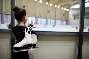 Back view of a young girl holding figure skates standing by an ice rink and watching others train