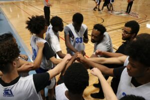 A coach with BIPOC basketball players in a huddle on the sidelines of a basketball court