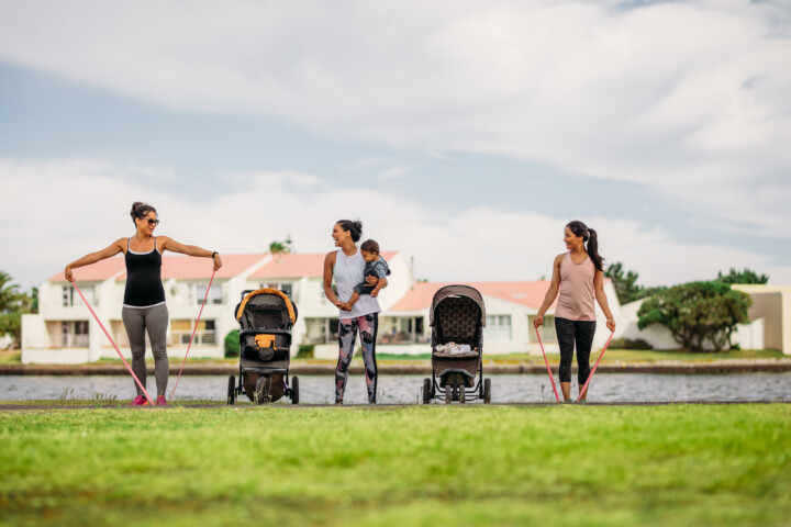 Women doing workout in park bringing their kids in strollers. Young mothers doing stretching exercise using stretch banks in park beside a lake.