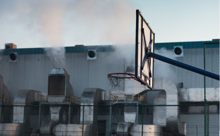 A basketball net with only a chainlink fence separating the court from a polluting industrial building