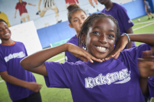 A young Black girl in a purple shirt smiles at the camera with hands framing her face