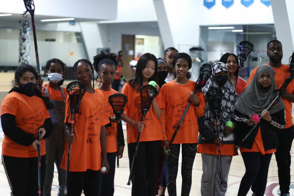 Youth athletes wearing orange shirts and holding lacrosse sticks