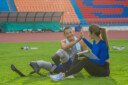 Athlete with prosthetics and trainer exchange a high-five on a grass lawn after a successful speed running practice at the stadium
