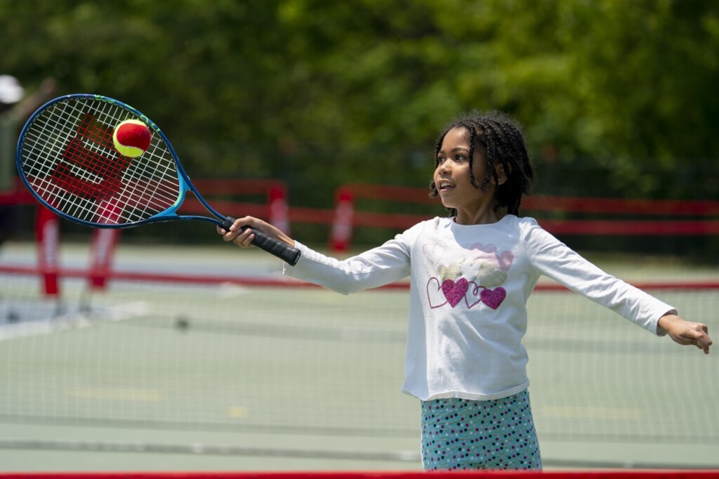 A child wearing a white, long-sleeved shirt and blue shorts is holding a tennis racket and hitting a tennis ball on a sunny day.