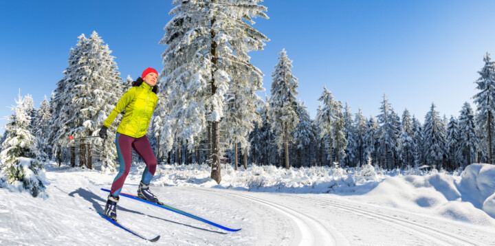 A woman with long brown hair, a red toque, yellow jacket and burgandy tights, cross country skis in the woods.