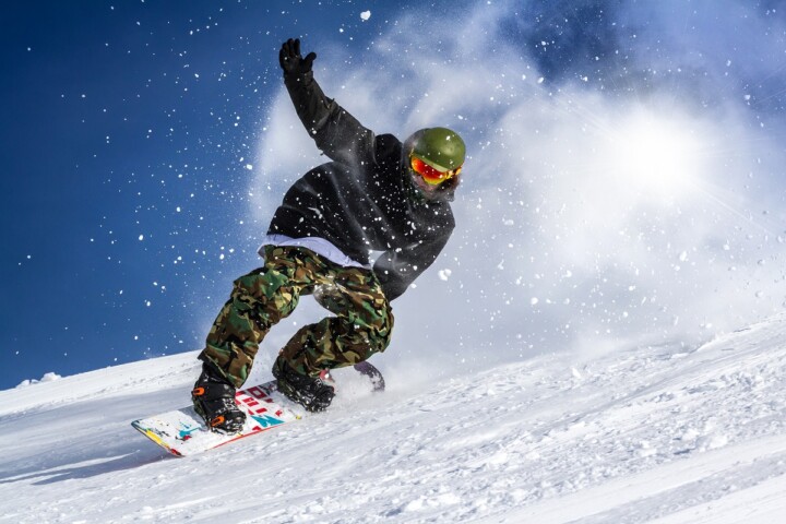 A young man with a helmet and goggles on is snowboarding down a mountain and has one hand in the air as snow flies.