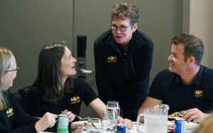 One woman stands in the middle of another man and woman, who are sitting at a table. 