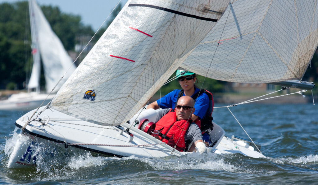 A closeup of a sailboat speeding towards the camera, spraying waves from its oncoming bow. A man (Bryan Cuerrier) with a red lifejacket, sunglasses, and a shaved head sits facing forward in the cockpit, intensely focused. There is a white and red windlass perched on the boat’s deck at chest level. The man’s left arm leans on the deck and is amputated at the forearm, and a young woman in a blue shirt and sunglasses is seated behind him.